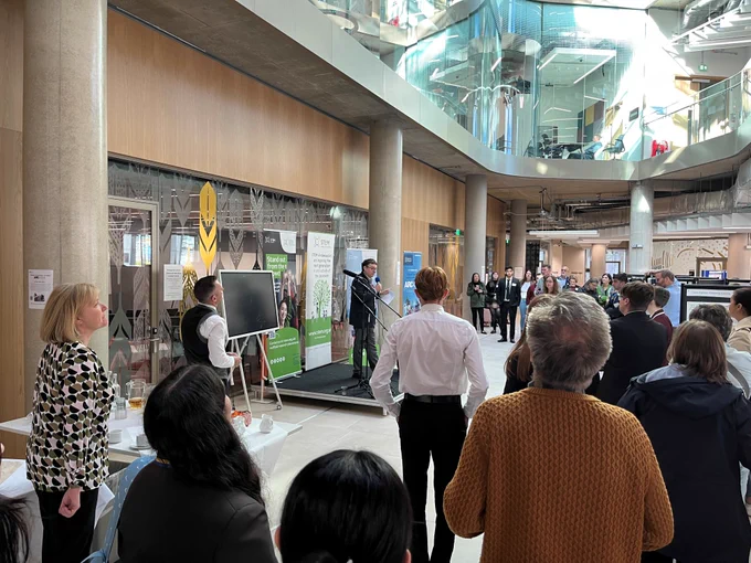A large crowd in a large glass atrium are looking at a speaker on a small stage