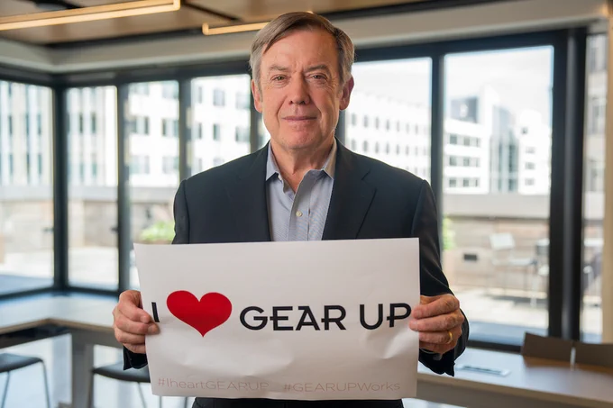 A person wearing a jacket and button down shirt smiles and holds up a sign that reads, "I (heart) Gear Up" in front of a bank of floor to ceiling windows.