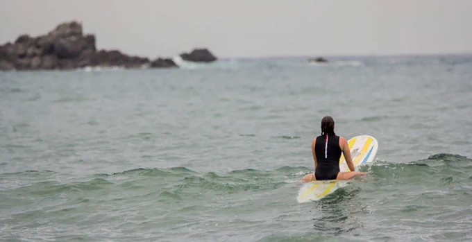 The Social Lights CEO Emily Pritchard paddling out to sea on a surfboard in the ocean with a rock cropping in the distance