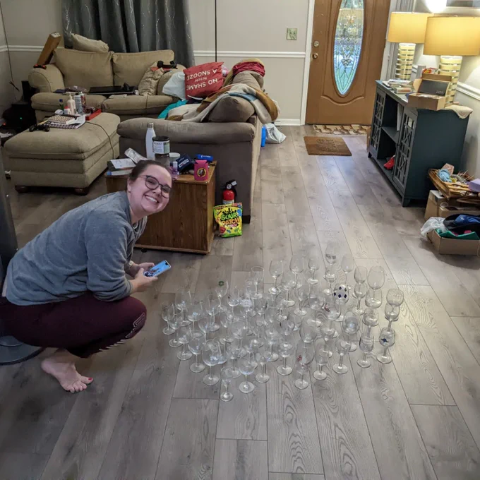 A white woman in a grey sweatshirt smiles as she squats next to a collection of 50 wine glasses. They are sitting on the floor in the middle of a living room.
