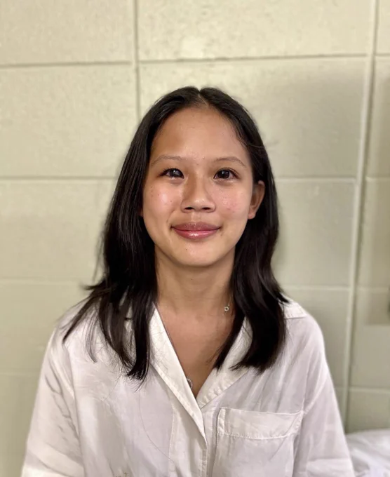This photo shows Malia sitting in front of a white, cinder lock wall. Malia is wearing a white button-up short-sleeve shirt with a pocket on one side. She has dark brown hair and dark brown eyes, and she is smiling at the camera.