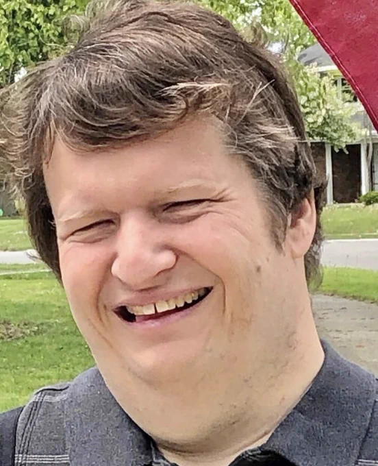 This photo shows Gregg standing from the shoulders up outside. He is wearing a dark grey polo shirt with thin white stripes on it, and he is smiling at the camera. Gregg has brown hair, and brown eyes.