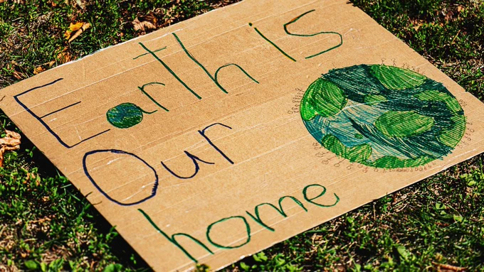 A piece of cardboard lying on top of grass, with an illustration of stick men around a blue-green Earth. The cardboard reads, Earth is our home.