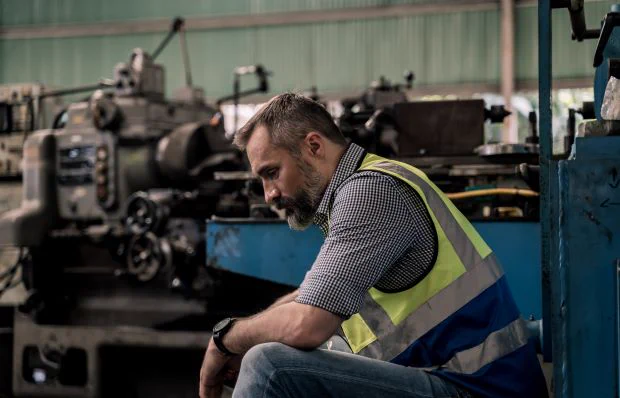 A greying bearded man in a high vis vest sits downcast in a factory with machinery in the background
