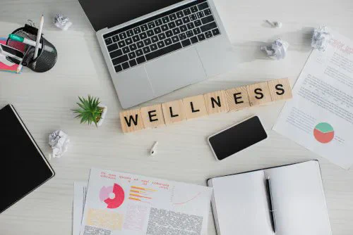 photo illustration that shows a desk with a laptop, pen cup, notebook, papers, and plants on it with the word Wellness spelled out in letter tiles