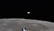 View of the Apollo 11 ascent module in the foreground flying above the grey Moon. Above the Moon’s horizon, the Earth is partly in the Moon’s shadow against the black of space (known as Earthrise).