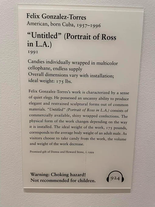 A banal beige Art Institute of Chicago gallery plaque reading:

Felix Gonzalez-Torres
American, born Cuba, 1957-1996

"Untitled" (Portrait of Ross in L.A)
1991

Candies individually wrapped in multicolor cellophane, endless supply
Overall dimensions vary with installation; ideal weight: 175 lbs. 

Felix Gonzalez-Torres's work is characterized by a sense of quiet elegy. He possessed an uncanny ability to produce elegant and restrained sculptural forms out of common materials. "Untitled" (Portrait of Ross in L.A.) consists of commercially available shiny wrapped confections. The physical form of the work changes depending on the way it is installed. The ideal weight of the work, 175 pounds, corresponds to the average body weight of an adult male. As visitors choose to take candy from the work, the volume and weight of the work decrease. 
