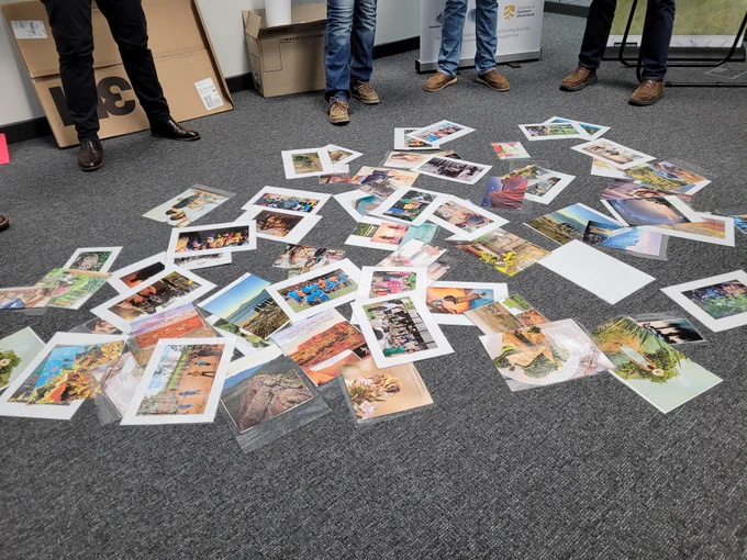 The photograph is of a group of people standing in a circle around printed photographs spread across the floor. All that can be seen of the people are four sets of legs.