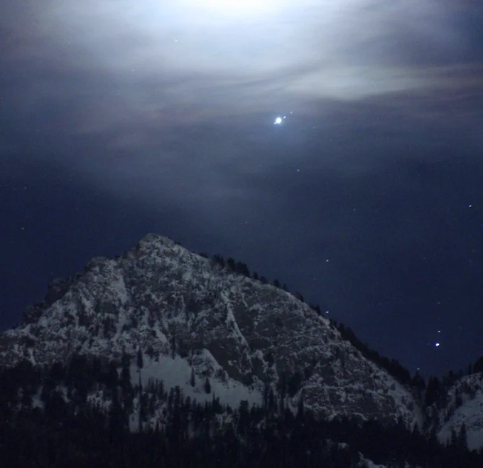 Jupiter is visible as a bright "star" in a partially cloudy night sky over a mountain range, along with three smaller stars right next to it, which are three of the planet's moons. Credits: NASA/Bill Dunford