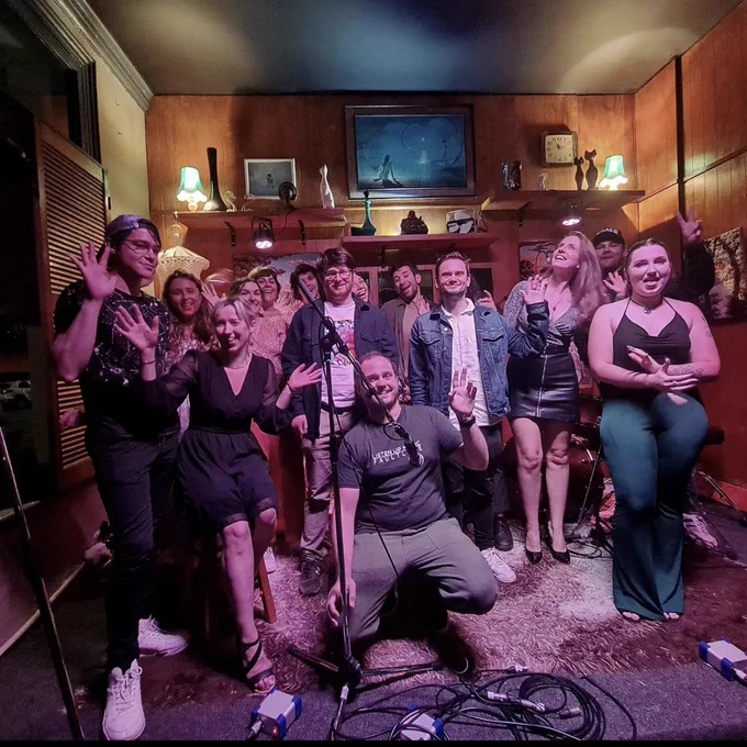 A group of songwriters and musicians smiling and waving on the intimate stage at Brisbane venue It’s Still a Secret.