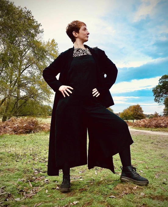 Ginger haired woman standing on some grass near trees, rocking a power pose in black dungarees. 
