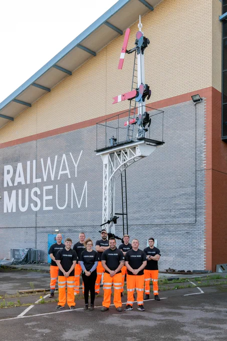 Portrait image showing colleagues from the NRM's Conservation team and Network Rail. They are all wearing black t-shirts that read 'Vision 2925' and the majority are wearing florescent orange work trousers. The somersault signal is directly behind them and the NRM logo is displayed on the wall behind the signal.
