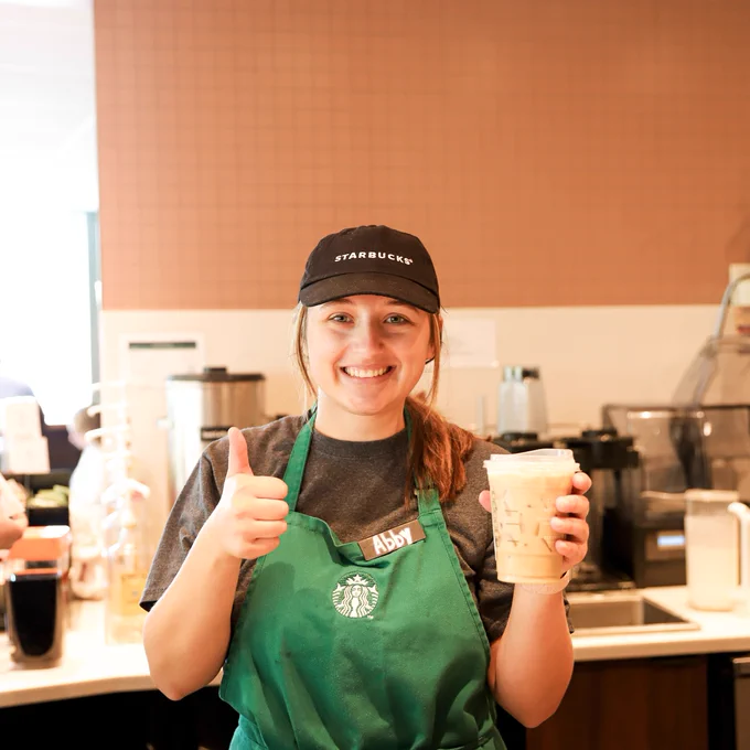 A woman wearing a green Starbucks apron is smiling at the camera. She is holding a iced coffee and giving a thumbs up.