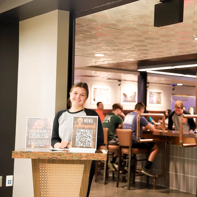 A woman stands at a counter and smiles at the camera. There are people in the bacckground sitting at a bar.