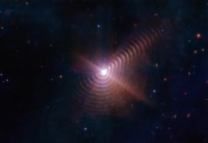 This Webb image shows a bright dot at the center of star-filled black space. The bright dot is actually two stars meeting, as their orbits bring them together every 8 years. The stellar pair are surrounded by 17 rings of gas and dust that appear orangish pink, like the rings of a tree trunk. The rings have a slight rectangular shape and are very clear and defined starting at about 1 o’clock on a clockface. The rings start to break up a bit to our view traveling clockwise around the image. As you arrive at the 8 o’clock position, only parts of about six rings can be seen as they disappear from view.