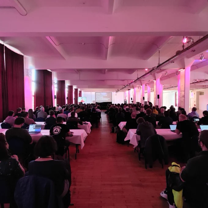 A photo taken at RustFest 2022 from the back of the audience. Two rows of seats and tables are leading up to the slide projection in the far end of the venue, with a smaller screen on the bottom left showing the live feed from the remote presenter's camera.