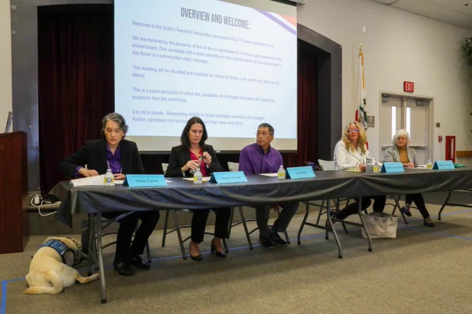 Candidates including Thida Cornes sit at a table debating. Thida is wearing a navy blue jacket and black pants. Her service dog wearing a blue vest lies at her feet.