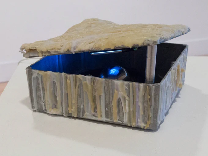 a metal box is dripped in wax to form a clamlike shape; its lid rests askew, offering a peek inside at a shiny metal orb, which is reflecting a small screen.