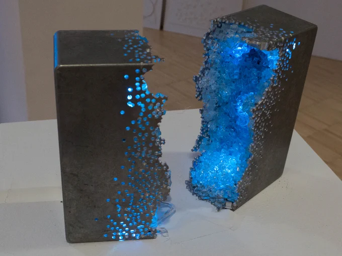 a metal box, split in half with hundreds of drilled holes, has a sparkling geode-like interior constructed with glowing glass "crystals." 
