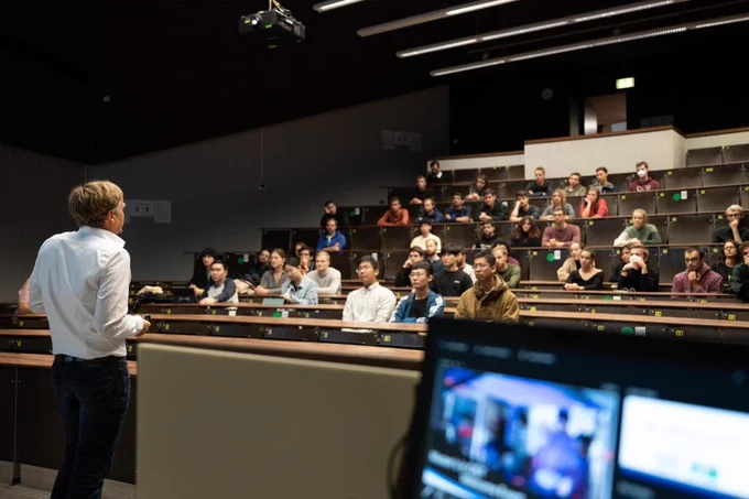 A lecture hall full of students with a speaker at the front, seen from behind the speaker. A laptop screen is in the foreground.