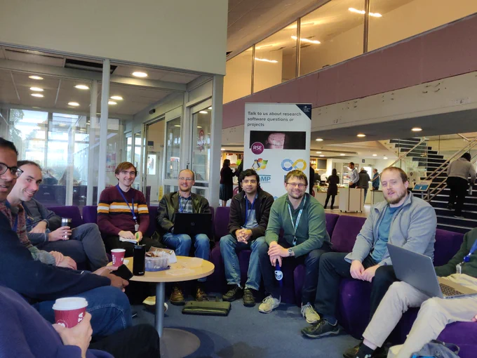 A group of research software engineers sitting in a semi-circle in front of a sign that says "Talk to us about research software questions or projects"