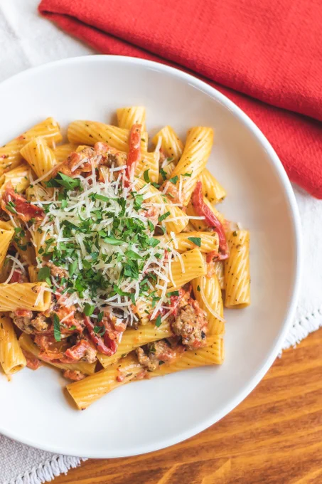 An overhead perspective of a white plate of pasta with grated cheese, sausage, and a tomato based sauce. The plate sits on a wooden table with a red napkin beside it.
