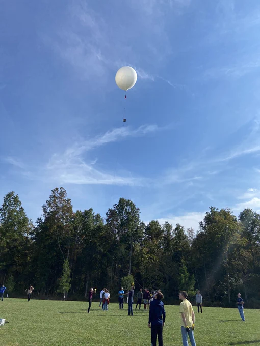A white weather balloon with an attached sensor container floats a hundred feet over a grass field. Students and teachers stand below. Two students hold tethers to control the balloon during this test flight.