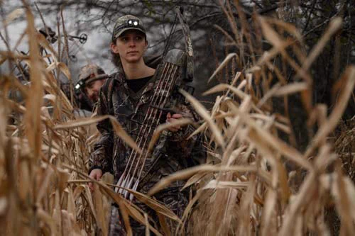 A female bowhunter dressed in camouflage and carrying a crossbow, walking through a cornfield with her hunting partner behind her.