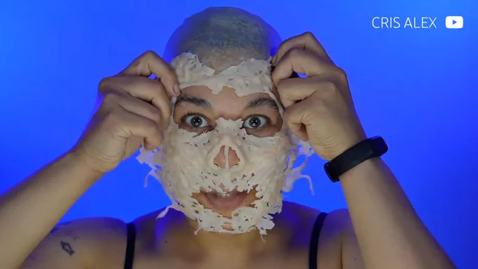 An image of creator Cris Alex in front of a blue background, holding a white prosthetic over their face. The prosthetic is skelental, extremely textured, and is missing a nose, while wearing a black halter top.