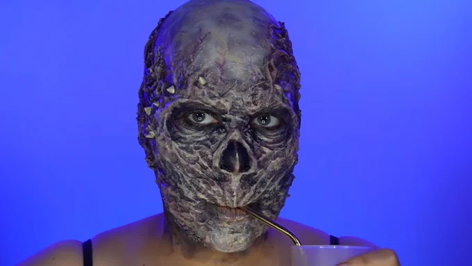 An image of creator Cris Alex in front of a blue background with a skeletal, extremely textured prosthetic that is missing a nose and features horns and veins, all painted a shiny black. Cris Alex wears a black tanktop and sips from a silver straw out of a frosted white cup. 