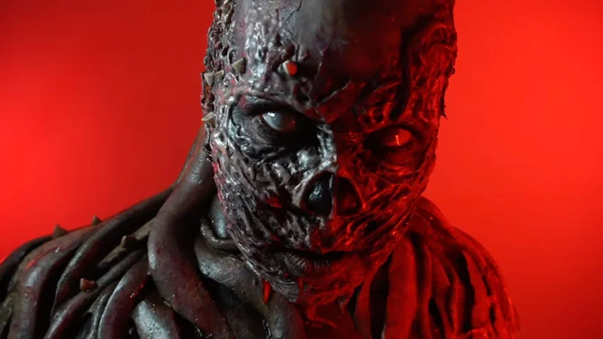 An image of Cris Alex in front of a red background revealing a full, dramatic Vecna from Stranger Things costume, which consists of a skeletal, extremely textured prosthetic that is missing a nose and features horns and veins, all painted a shiny black with bulbous flesh hanging from the next.