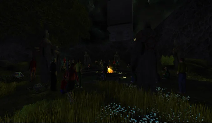 A large group of players gathered around a pond in a lush overgrown space at dusk. In the centre of the pond, a large yellow ornamental lamp casts some light. Surrounding the pond are large stone statues. Long grass and small blue flowers wave slightly in a gentle breeze.