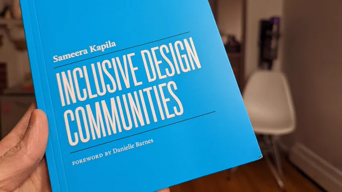 Hand holding book who is cover reads inclusive design communities. In the background is a white chair.