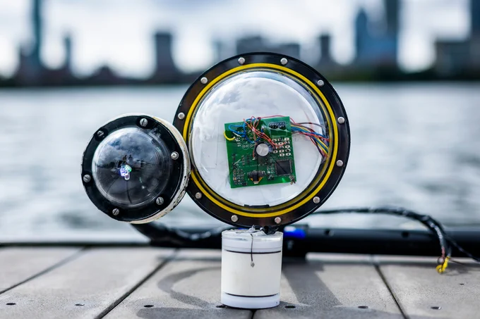 A prototype camera, with 2 bulbous lenses, a circuit board, wires and white cylinder, sits on a dock with the Charles River and Boston skyline in blurred background.