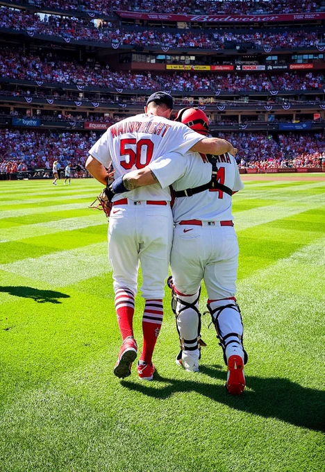 Adam Wainwright and Yadier Molina walk out from the bullpen to the home dugout with their arms around each other. Wainwright's right arm is over Molina's shoulders and Molina's left arm is around Wainwright's back.