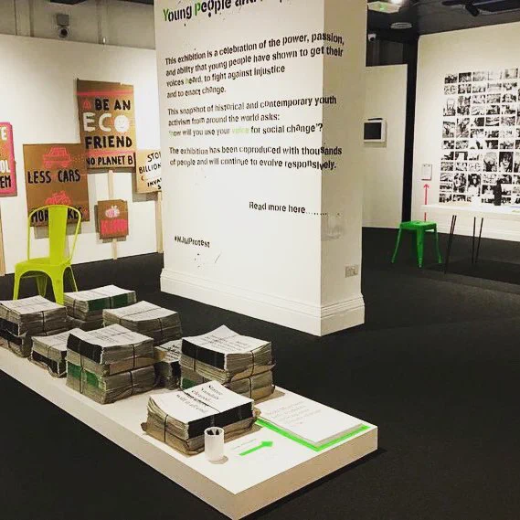 a fallers with black floor  white walls, text wall, black and white protest photos, and bright pops of fluro tape on card placards
