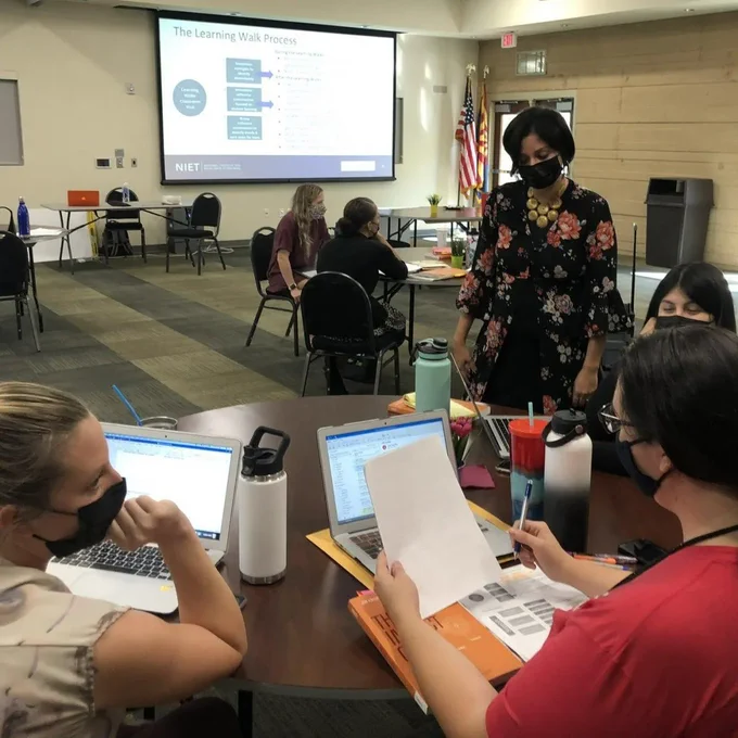A teacher leading a professional learning session in a large meeting space with a presentation projected on the screen as they interact with participants at tables