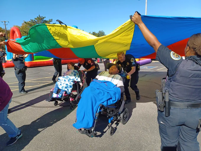 Two officers push students using wheelchairs under a rainbow-colored parachute held up by other officers.