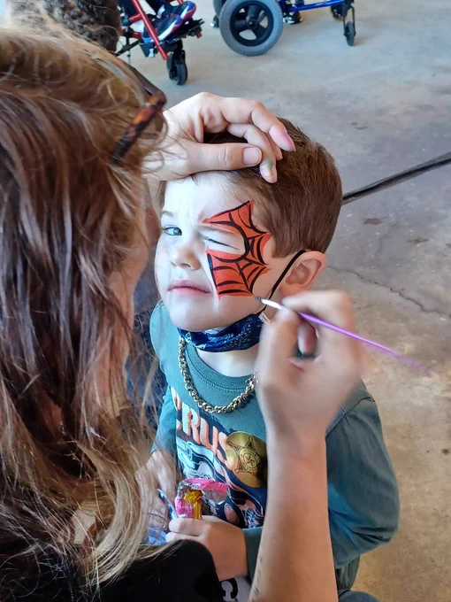 Child having face painted with an orange and black cobweb.