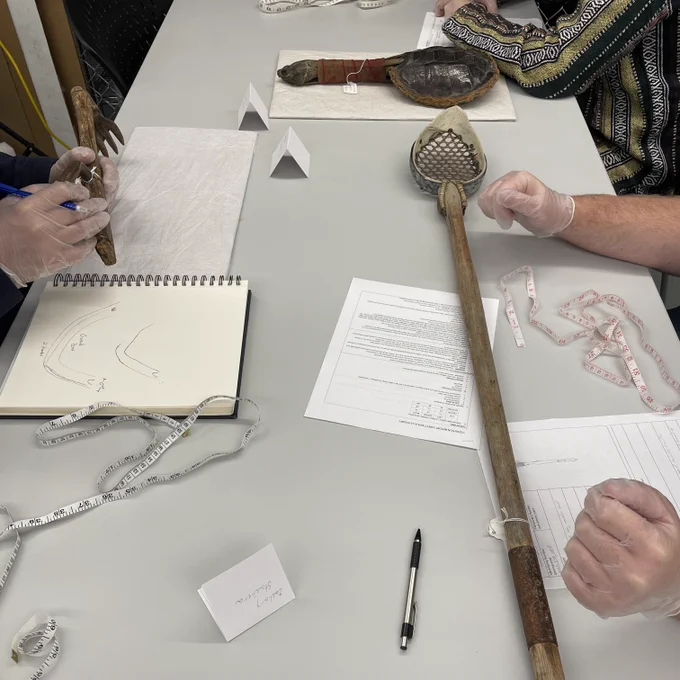 A variety of artifacts, including instruments and weapons, arranged on a table along with sheets of paper, pencils, notecards, and a notebook. Gloved hands are lifting up the objects and handling them.