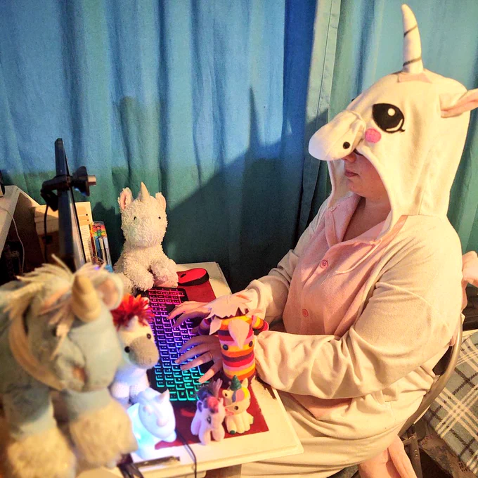 A woman in a white unicorn onesie sat typing at a desk surrounded by various toy unicorns