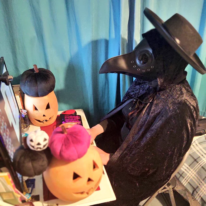 A woman sat at a desk dressed all in black, wearing a black cloak, hat and plague doctor mask, surrounded by pumpkins and jack o'lanterns