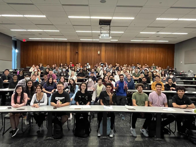 Teaching assistants pose for a picture inside a brightly-lit lecture hall.

The picture is taken from the front of the room; there are around seventy TAs, seated and smiling, a handful of them with their hands raised as if to wave. The tables are horizontally long and cascade upwards, leading one's eye to the brown wall at the back of the room. 