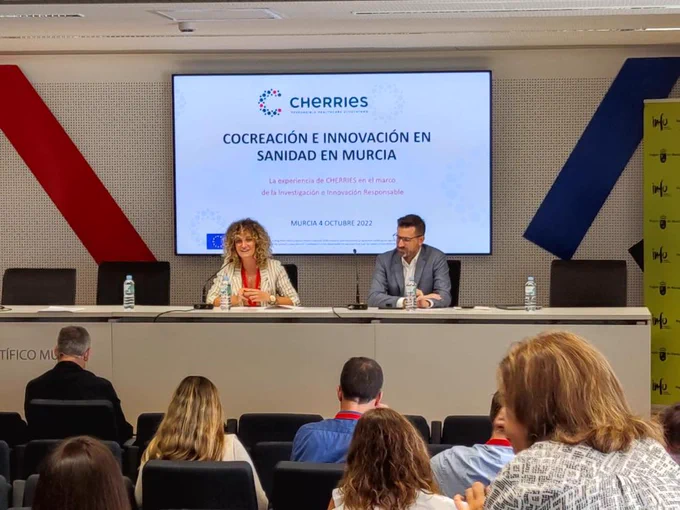 A woman and a man on a stage introducing the event " Cocreation and innovation in the healthcare sector in Murcia" in front of an audience sitting in the room.