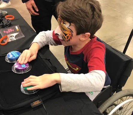 A picture of a child in a wheelchair playing a videogame with very large buttons in front of them, tiger face paint on their face, staring intensely at the screen, ablegamers pamphlets and bracelets and switches visible on the table in the background