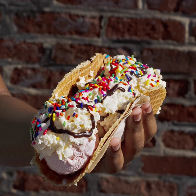 A hand holds an ice cream taco with scoops of ice cream topped with whipped cream, hot fudge and rainbow sprinkles. A brick wall is the background
