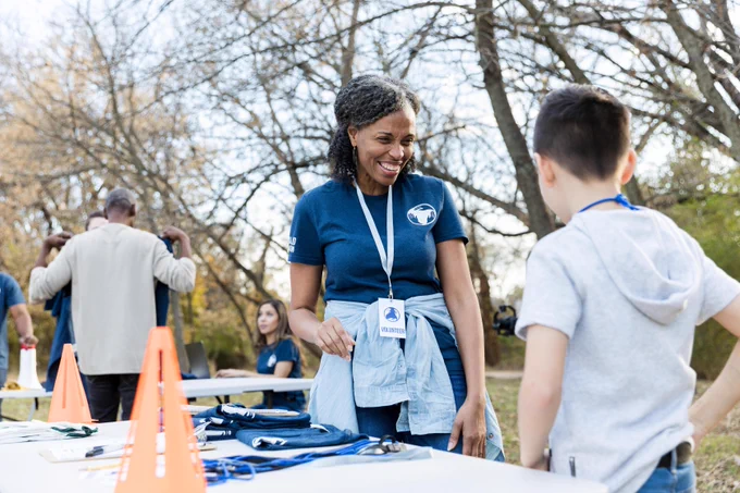 A woman in a blue shirt talks with a young boy with his back to the camera. They stand by a table. 