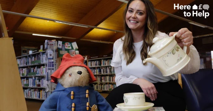 A woman wearing a white top and black trousers is pictured sat next to a Paddington Bear teddy, on chair in Perton Library. The woman is pretending to pour tea from a white teapot into a white cup and saucer. A white Here to Help logo is in the top right hand corner of the image.