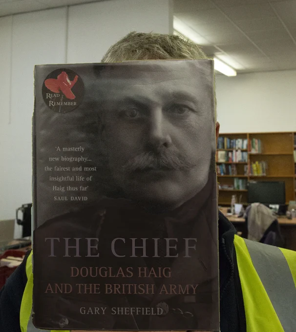 Image of a person in front of a shelf of books and a computer screen holding the biography titled The Chief Douglas Haig and the British Army by Gary Sheffield in front of their face.