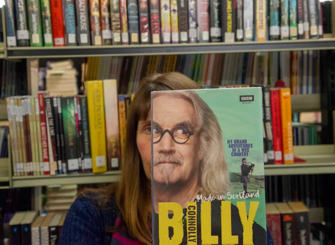 Image of a person sitting in front of a shelf of books holding Billy  Connolly's autobiography, Made in Scotland, in front of their face.